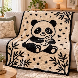 May include: A cream-colored crochet blanket featuring a large black and white panda bear holding a baby bottle. The design includes black bamboo stalks in the corners, scattered black stars, and small black hearts across the surface.