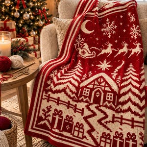 May include: A red and cream knit throw blanket featuring a festive holiday pattern with a central cabin, pine trees, a sleigh with reindeer, snowflakes, a crescent moon, and gift boxes along the bottom border, draped over a textured armchair.