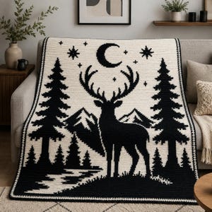 Peut inclure: Une couverture en crochet rectangulaire pr&eacute;sentant un motif contrast&eacute; noir et cr&egrave;me. Le design repr&eacute;sente la silhouette centrale d'un cerf aux grands bois, entour&eacute; de sapins, de montagnes, d'un croissant de lune et de petites &eacute;toiles.