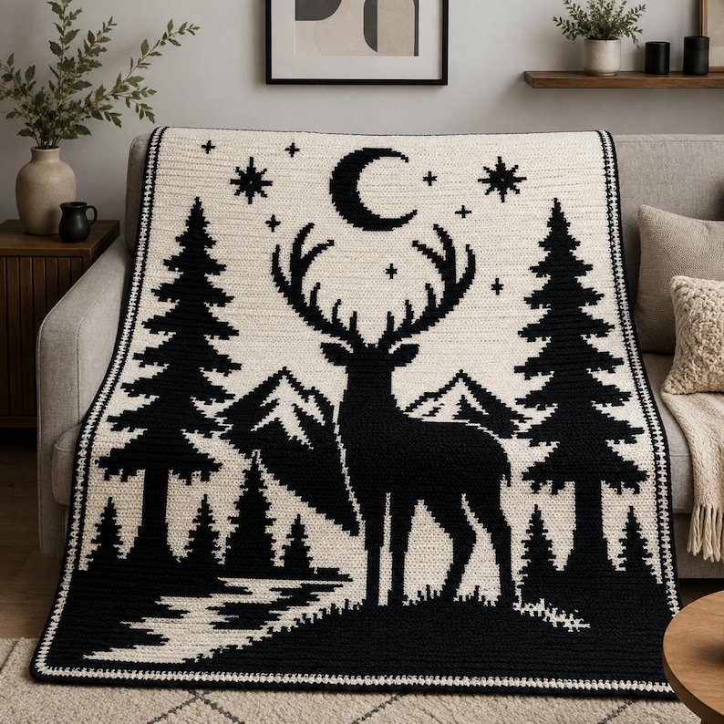 Peut inclure: Une couverture en crochet rectangulaire pr&eacute;sentant un motif contrast&eacute; noir et cr&egrave;me. Le design repr&eacute;sente la silhouette centrale d'un cerf aux grands bois, entour&eacute; de sapins, de montagnes, d'un croissant de lune et de petites &eacute;toiles.
