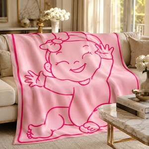 Pu&ograve; includere: Una coperta rosa con una grande grafica di un bambino sorridente in un contorno rosa pi&ugrave; scuro. La coperta &egrave; drappeggiata su un divano beige. L'illustrazione del bambino ha un fiocco e agita le mani. La coperta ha un bordo rosa pi&ugrave; scuro.
