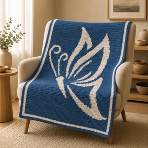 May include: A blue and white crochet blanket with a butterfly design, draped over a chair. The blanket has a white butterfly on a blue background, with a white border. The chair is beige, and the background includes a window and wooden shelves.