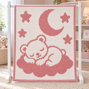 May include: A rectangular crochet blanket featuring a cream-colored background with a dusty rose pink border. The design displays a sleeping bear resting on a cloud, surrounded by a crescent moon and three stars, all rendered in a matching rose pink.