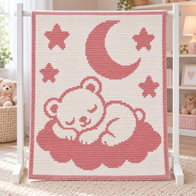May include: A rectangular crochet blanket featuring a cream-colored background with a dusty rose pink border. The design displays a sleeping bear resting on a cloud, surrounded by a crescent moon and three stars, all rendered in a matching rose pink.