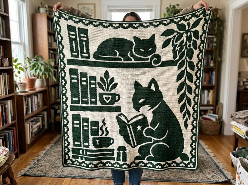May include: A cream-colored crochet blanket featuring a dark green design of two cats, bookshelves, potted plants, and a steaming mug. The border consists of a repeating scalloped pattern in dark green and cream, framing the entire rectangular piece.