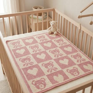 May include: A pink and cream-colored baby blanket with a teddy bear and heart pattern. The blanket is placed in a wooden crib with a stuffed teddy bear. The blanket is made of a soft material and is likely handmade.