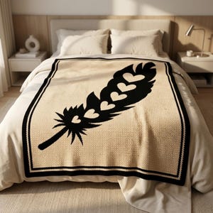 May include: A cream-coloured knitted blanket with a large black feather design featuring heart-shaped cutouts. The blanket has a black border and is displayed on a bed with white bedding. The overall theme is home decor.