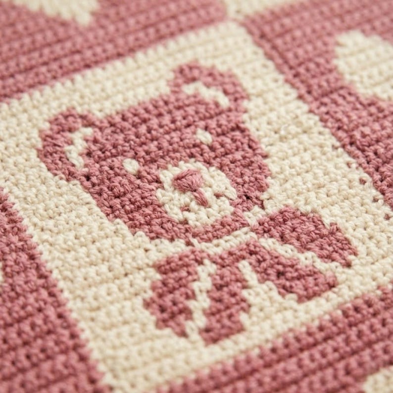 May include: Close-up of a handmade crochet blanket featuring a repeating pattern of teddy bears in a soft pink color against a cream background. The blanket is made with a tight stitch, creating a textured surface.