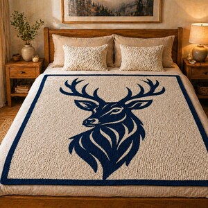 May include: A beige and navy blue knitted throw blanket featuring a large navy blue deer silhouette. The blanket has a navy blue border and is displayed on a bed with matching pillows and wooden furniture. The overall theme is rustic and outdoorsy.