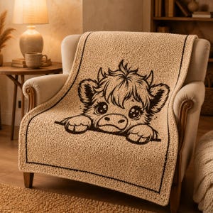 May include: A beige throw blanket featuring a cartoon cow face design with a dark brown border. The blanket is draped over a cream armchair. The cow has large eyes and is peeking over the edge of the blanket.