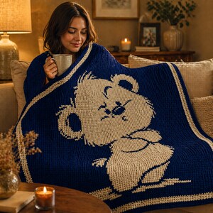 May include: A navy blue knitted blanket with a cream-coloured koala bear design. The blanket has a cream border and is draped over a person. This soft blanket is a warm home decor item, perfect for snuggling up on the sofa.