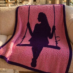 Puede incluir: Una manta rosa tejida a ganchillo con una silueta p&uacute;rpura oscura de una persona en un columpio. La manta tiene un borde p&uacute;rpura oscuro. La manta est&aacute; sobre un sof&aacute; beige.
