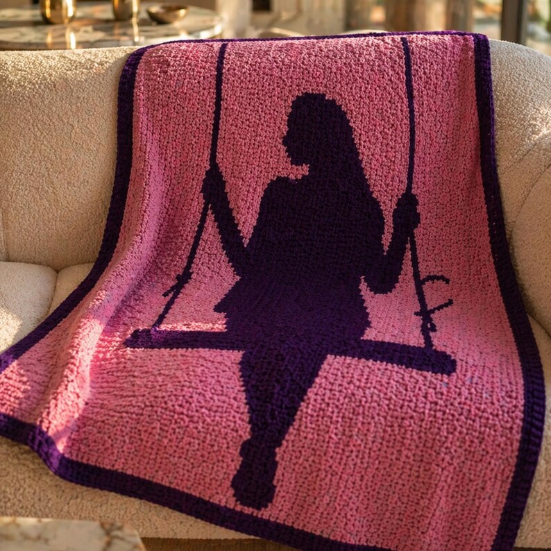 Puede incluir: Una manta rosa tejida a ganchillo con una silueta p&uacute;rpura oscura de una persona en un columpio. La manta tiene un borde p&uacute;rpura oscuro. La manta est&aacute; sobre un sof&aacute; beige.