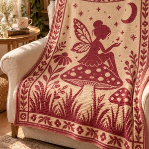 May include: A cream-colored crochet blanket featuring a dark red pattern of a fairy sitting on a large mushroom, surrounded by smaller mushrooms, flowers, and a crescent moon with stars, all enclosed within a decorative floral border.