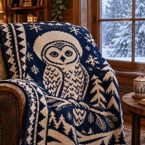May include: A navy blue and cream-colored knit throw blanket featuring a large owl wearing a hat, surrounded by snowflake and pine tree patterns. The blanket is draped over a brown leather chair, showcasing a detailed winter-themed design.