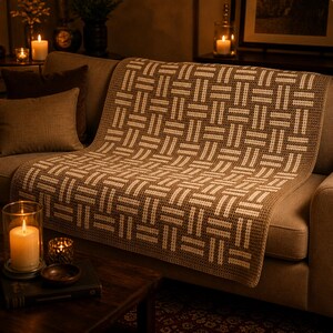 May include: A brown and cream-colored crocheted blanket with a geometric pattern is draped over a sofa. The blanket's design features a repeating woven-like motif. The sofa has two throw pillows and is in a cozy living room setting.