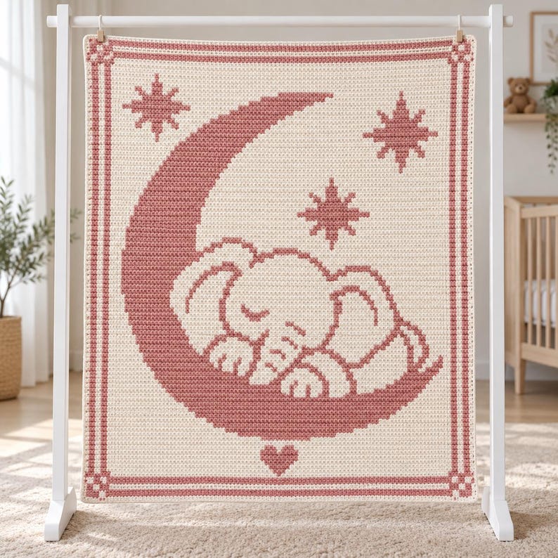 May include: A rectangular crochet blanket featuring a cream-colored base with a dusty rose pattern. The design displays a sleeping elephant resting on a crescent moon, surrounded by three eight-pointed stars and a small heart at the bottom center.