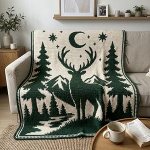 Peut inclure: Une couverture en crochet de couleur cr&egrave;me pr&eacute;sentant une sc&egrave;ne de for&ecirc;t vert fonc&eacute; avec une silhouette de cerf au centre, des montagnes, des sapins, un croissant de lune et des &eacute;toiles. Le motif est bord&eacute; d'une bordure g&eacute;om&eacute;trique.