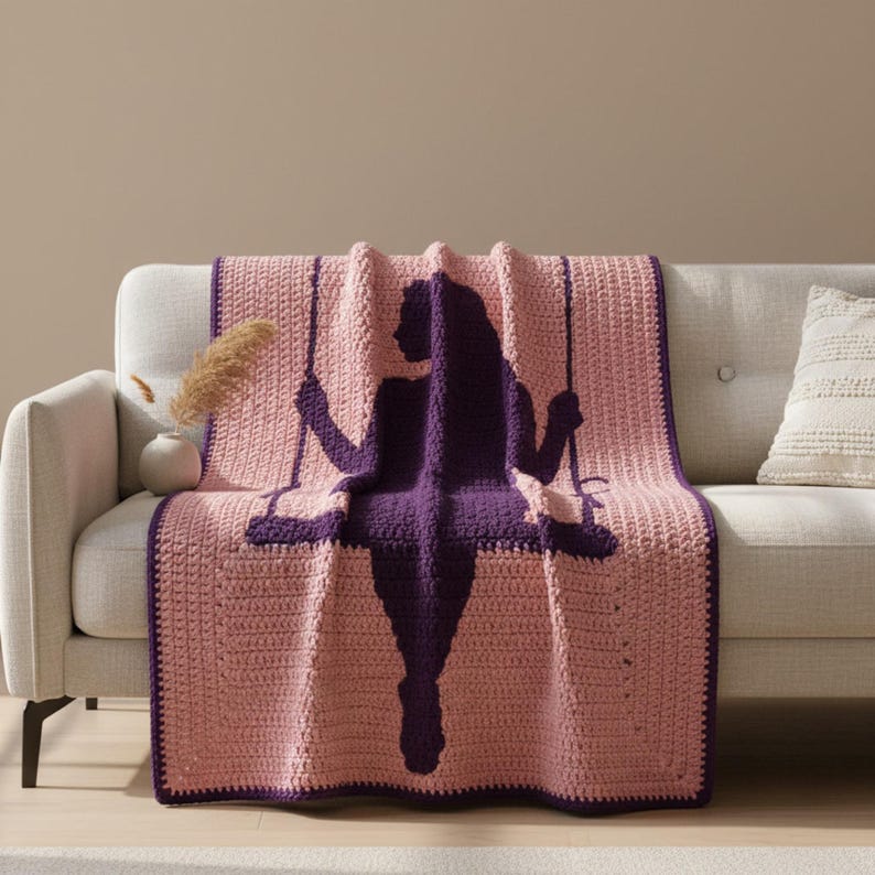 Puede incluir: Una manta de ganchillo en tonos rosa y morado, colocada sobre un sof&aacute;. La manta presenta la silueta de una persona en un columpio en un tono morado m&aacute;s oscuro. Los bordes de la manta est&aacute;n ribeteados con un borde morado a juego.