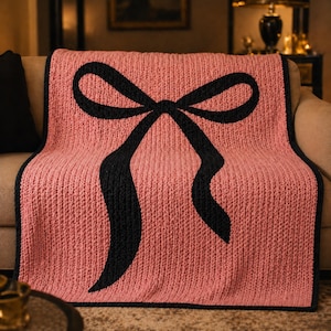 Pu&ograve; includere: Una coperta rosa lavorata a maglia con un grande disegno a fiocco nero. La coperta ha una trama a coste e un bordo nero. Il fiocco &egrave; centrato sulla coperta, con le estremit&agrave; del nastro che scorrono verso il basso. La coperta &egrave; drappeggiata su un divano.