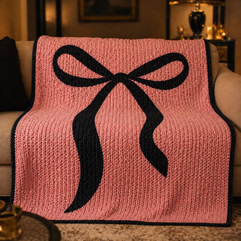 Pu&ograve; includere: Una coperta rosa lavorata a maglia con un grande disegno a fiocco nero. La coperta ha una trama a coste e un bordo nero. Il fiocco &egrave; centrato sulla coperta, con le estremit&agrave; del nastro che scorrono verso il basso. La coperta &egrave; drappeggiata su un divano.