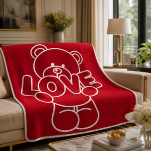 May include: A red throw blanket with a white outline of a teddy bear holding the word "LOVE". The blanket has a white border and is draped over a beige sofa. The design is simple and modern.