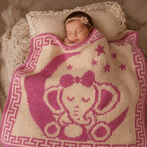 May include: A sleeping baby wrapped in a pink and cream-colored knitted blanket. The blanket features a pink elephant design with a bow, stars, and a Greek key border. The baby wears a floral headband.