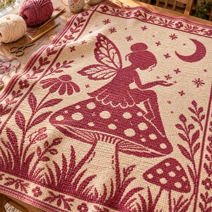 May include: A cream-colored crochet blanket featuring a dark red pattern of a fairy sitting on a large mushroom, surrounded by smaller mushrooms, flowers, and a crescent moon. The blanket has a decorative border with floral and leaf motifs.