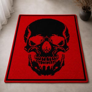 Puede incluir: Una manta de ganchillo roja y negra con un dise&ntilde;o de calavera. La calavera negra est&aacute; centrada sobre el fondo rojo. La manta tiene un borde negro. Los rasgos de la calavera son detallados, incluyendo cuencas de los ojos, nariz y dientes.