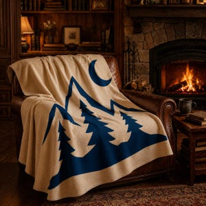 May include: A beige throw blanket draped over a brown leather armchair. The blanket features a dark blue graphic print of a mountain range with pine trees and a crescent moon positioned above the peaks, creating a high-contrast visual design.