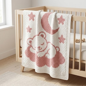 May include: A cream-colored knit blanket featuring a dusty rose pattern of a sleeping bear resting on a cloud, surrounded by a crescent moon and stars. The blanket has a textured grid-like weave and a solid border with a small square label.