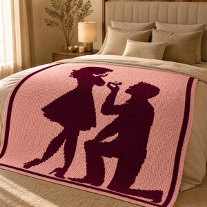 Pode incluir: Um cobertor rosa tricotado com uma borda bord&ocirc; e um design de silhueta de uma pessoa pedindo outra em casamento. O cobertor est&aacute; em uma cama com roupa de cama de cor neutra.