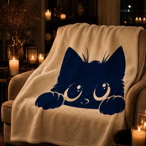 May include: A cream-colored sherpa throw blanket draped over a chair, featuring a navy blue cat face design. The cat has large, expressive eyes and paws. The blanket is in a cozy setting with candles and soft lighting.