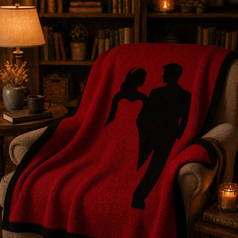 Pode incluir: Uma manta vermelha com uma borda preta e uma silhueta de um casal. A manta est&aacute; sobre uma cadeira em um ambiente aconchegante com uma l&acirc;mpada, livros e uma vela. O design da manta apresenta uma noiva e um noivo.