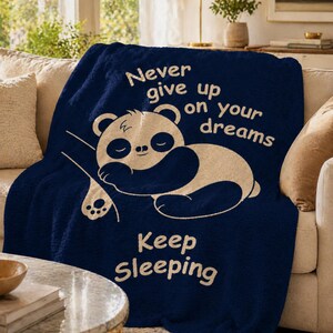May include: A navy blue throw blanket featuring a sleeping panda bear design. The blanket has the text "Never give up on your dreams" and "Keep Sleeping" in cream. The blanket is draped over a beige sofa.