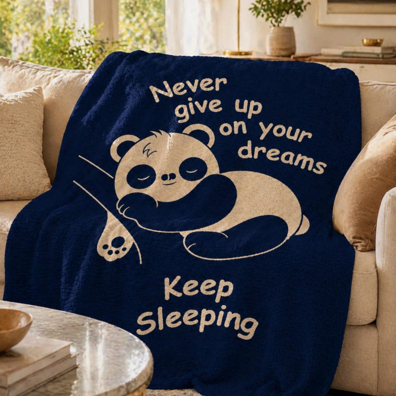 May include: A navy blue throw blanket featuring a sleeping panda bear design. The blanket has the text "Never give up on your dreams" and "Keep Sleeping" in cream. The blanket is draped over a beige sofa.