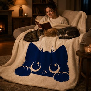 May include: A cozy cream-colored throw blanket with a navy blue cat face design. The blanket is draped over a chair, with three cats resting on it. A person is sitting in the chair, reading a book.