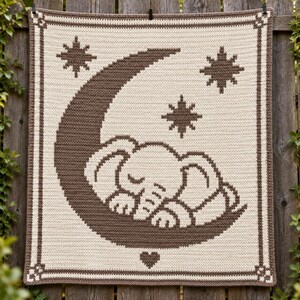 May include: A rectangular crochet blanket featuring a cream-colored background with a dark brown pattern. The design displays a sleeping elephant resting on a crescent moon, surrounded by three eight-pointed stars and a small heart at the bottom.