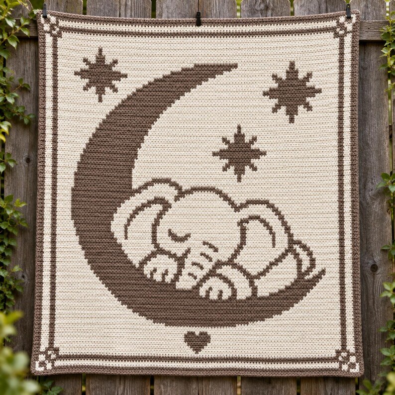 May include: A rectangular crochet blanket featuring a cream-colored background with a dark brown pattern. The design displays a sleeping elephant resting on a crescent moon, surrounded by three eight-pointed stars and a small heart at the bottom.