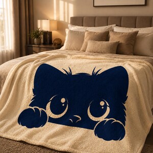 May include: A cream-colored sherpa blanket featuring a navy blue cat face design. The cat has large, round eyes and paws, creating a playful and cozy aesthetic. This blanket is designed for comfort and adds a touch of whimsy to any bedroom.