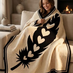 May include: Cream crochet blanket featuring a black feather design with white hearts. The blanket has a black border and is draped over a person on a beige sofa. A cosy home decor item, perfect for adding warmth and style.