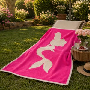May include: A bright pink beach towel with a white mermaid silhouette design. The towel is rectangular, with a white border, and is draped over a lounge chair on a grassy lawn. A straw hat and a small table with drinks and flowers are nearby.