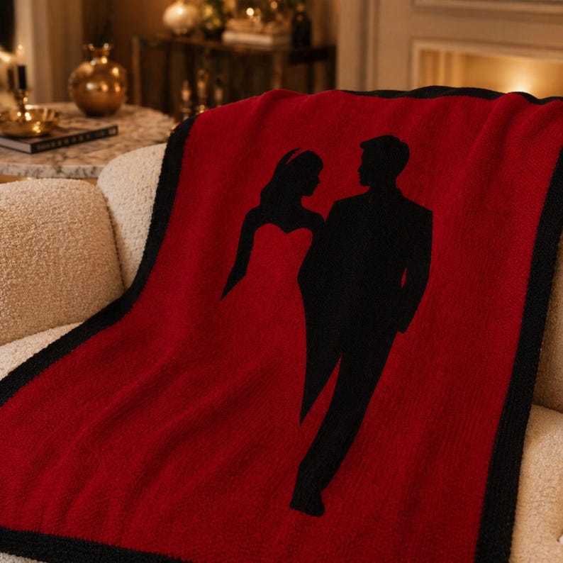 Pode incluir: Uma manta vermelha com uma borda preta e um design de silhueta de um casal. A mulher est&aacute; vestida com um vestido e o homem com um fato. A manta est&aacute; sobre um sof&aacute; creme.