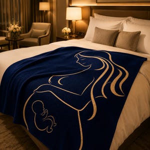 May include: A navy blue blanket featuring a gold illustration of a pregnant person and a fetus. The blanket is draped over a bed with white bedding and brown cushions. The room has warm lighting and a comfortable feel.