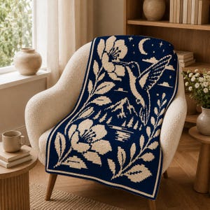 May include: A rectangular woven throw blanket featuring a dark navy blue background with a cream-colored graphic design. The pattern depicts a hummingbird hovering near a large flower, with mountains, a lake, trees, stars, and a crescent moon.