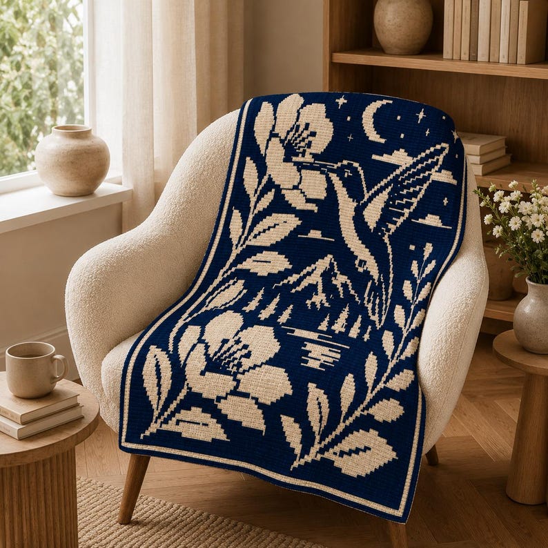 May include: A rectangular woven throw blanket featuring a dark navy blue background with a cream-colored graphic design. The pattern depicts a hummingbird hovering near a large flower, with mountains, a lake, trees, stars, and a crescent moon.