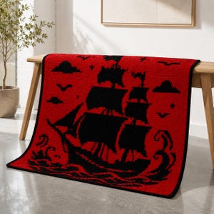 May include: A rectangular throw blanket featuring a vibrant red background with a black pixelated graphic of a sailing ship on the ocean. The design includes black silhouettes of clouds, birds, and stars surrounding the ship, with a black border.