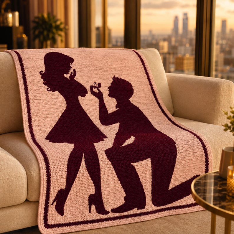 Pode incluir: Um cobertor rosa de croch&ecirc; com a silhueta de uma cena de pedido de casamento. O design mostra um homem ajoelhado oferecendo um anel a uma mulher. O cobertor tem uma borda bord&ocirc; escura e est&aacute; sobre um sof&aacute; bege.