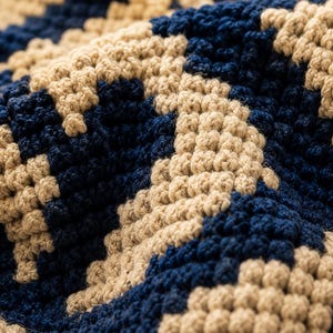 May include: Close-up of a textured, hand-crocheted fabric. The pattern features a repeating geometric design in navy blue and beige. The yarn appears thick and the stitches are tightly woven, creating a dense, tactile surface. The overall effect is of a cosy, handcrafted textile.