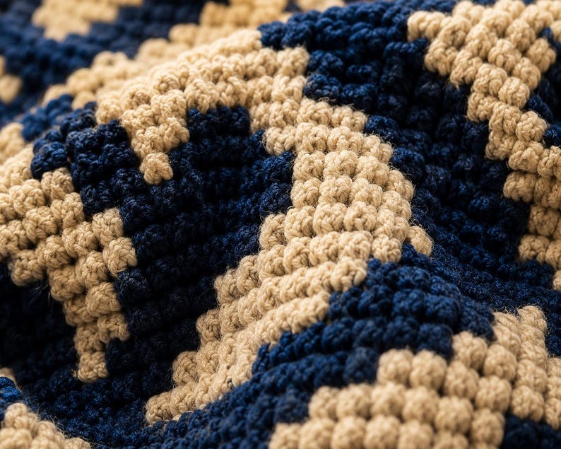 May include: Close-up of a textured, hand-crocheted fabric. The pattern features a repeating geometric design in navy blue and beige. The yarn appears thick and the stitches are tightly woven, creating a dense, tactile surface. The overall effect is of a cosy, handcrafted textile.